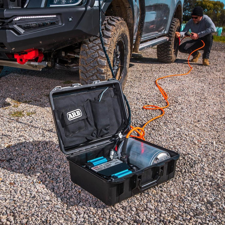 ARB CKMTP12 twin motor air compressor with ARB logo on a gravel surface, connected to a vehicle by an orange air hose.