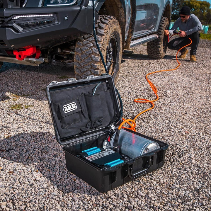 ARB CKMTP12 twin motor air compressor with ARB logo on a gravel surface, connected to a vehicle by an orange air hose.